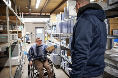 Foto toont persoon met handicap in rolstoel. In magazijn geeft hij een pakket af aan een collega.